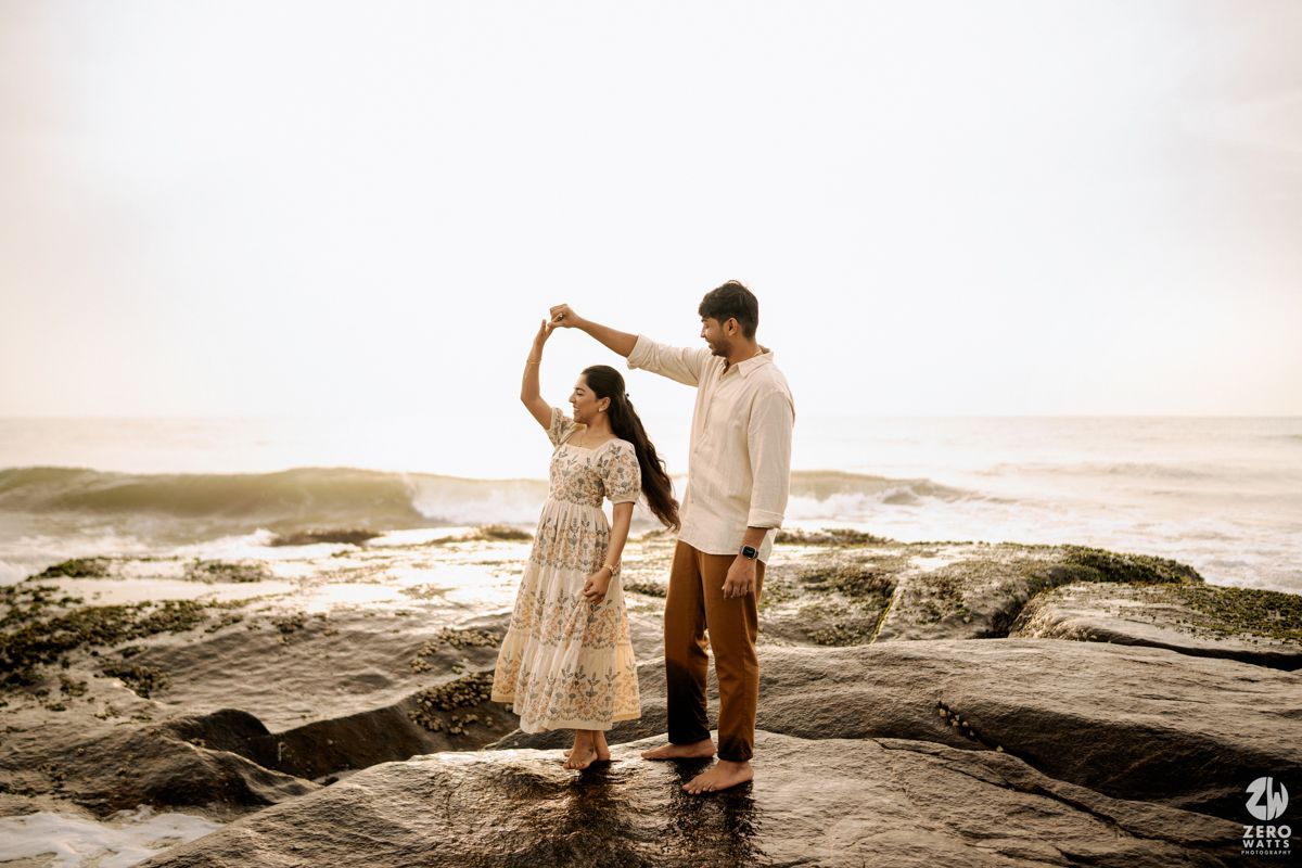 Couple walking along a rocky beach shoreline during golden hour in casual white outfits.