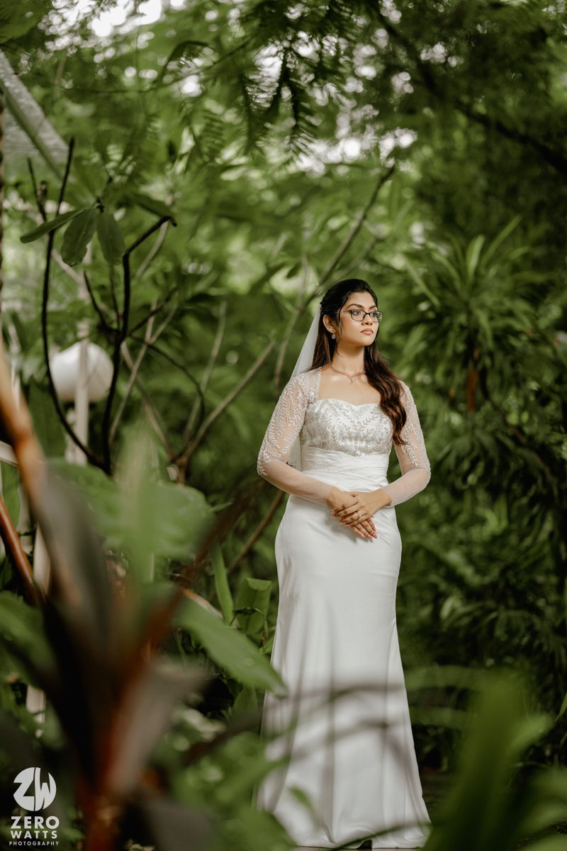 A bride in a white lace wedding dress poses in a lush tropical garden setting wearing glasses.