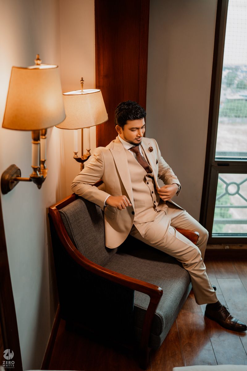 A person in a beige suit jacket and gray pants poses elegantly in an armchair near a window with warm lighting.