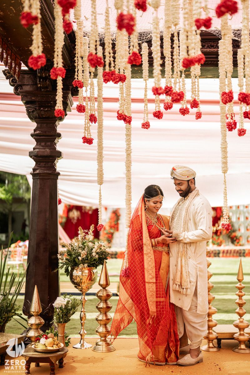 South Indian bride and groom standing under floral mandap decorated with red and white flowers.