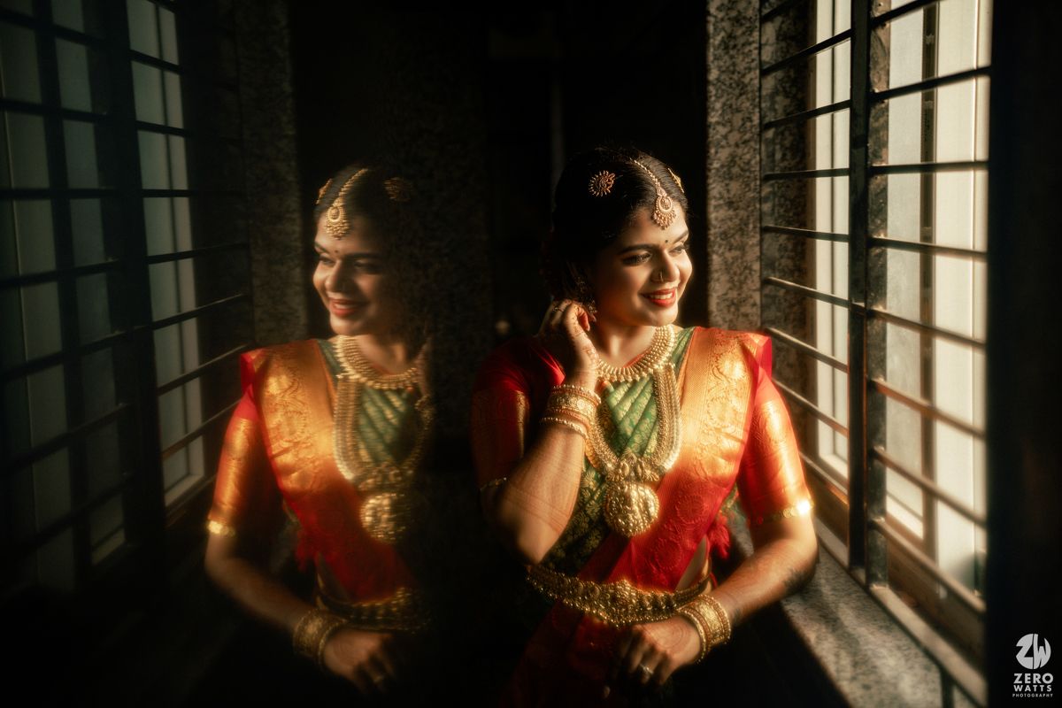South Indian bride in red saree and gold jewelry smiles near window, reflected in mirror during bridal photoshoot.
