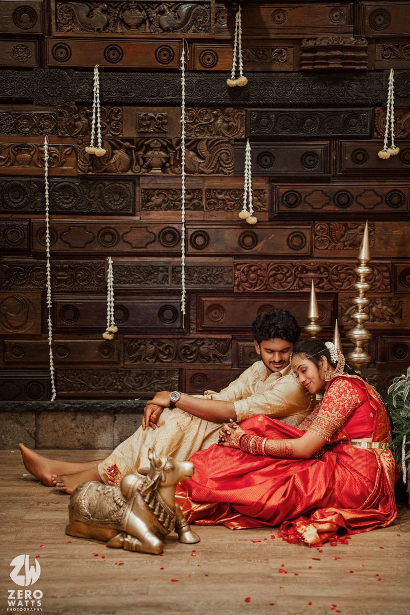 Indian bride in red saree and groom in cream outfit sitting together against ornate carved wooden backdrop.