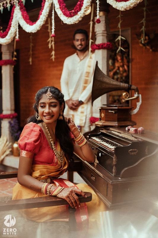 Smiling Indian bride in orange saree plays harmonium with groom in white dhoti standing behind near a vintage gramophone.