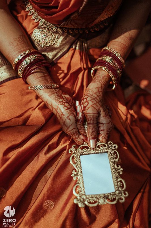 Bridal hands adorned with intricate mehndi designs, red bangles, and gold jewelry holding a ornate mirror in an orange saree.
