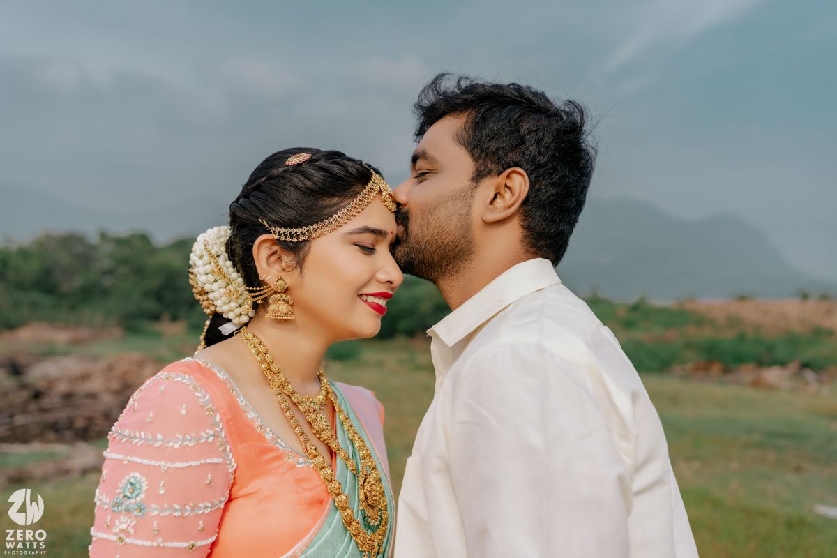 Indian bride and groom share a romantic moment outdoors, with mountains in the background, bride in pink saree and gold jewelry.