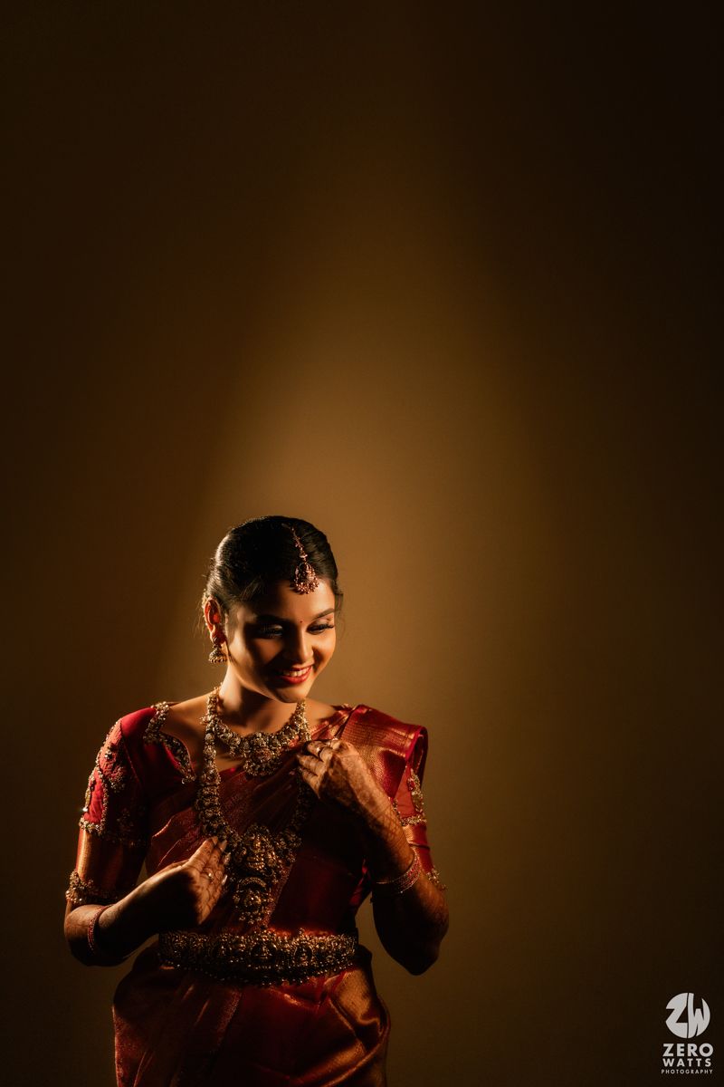 South Indian bride in red saree and gold jewelry smiles gracefully against a warm golden background.