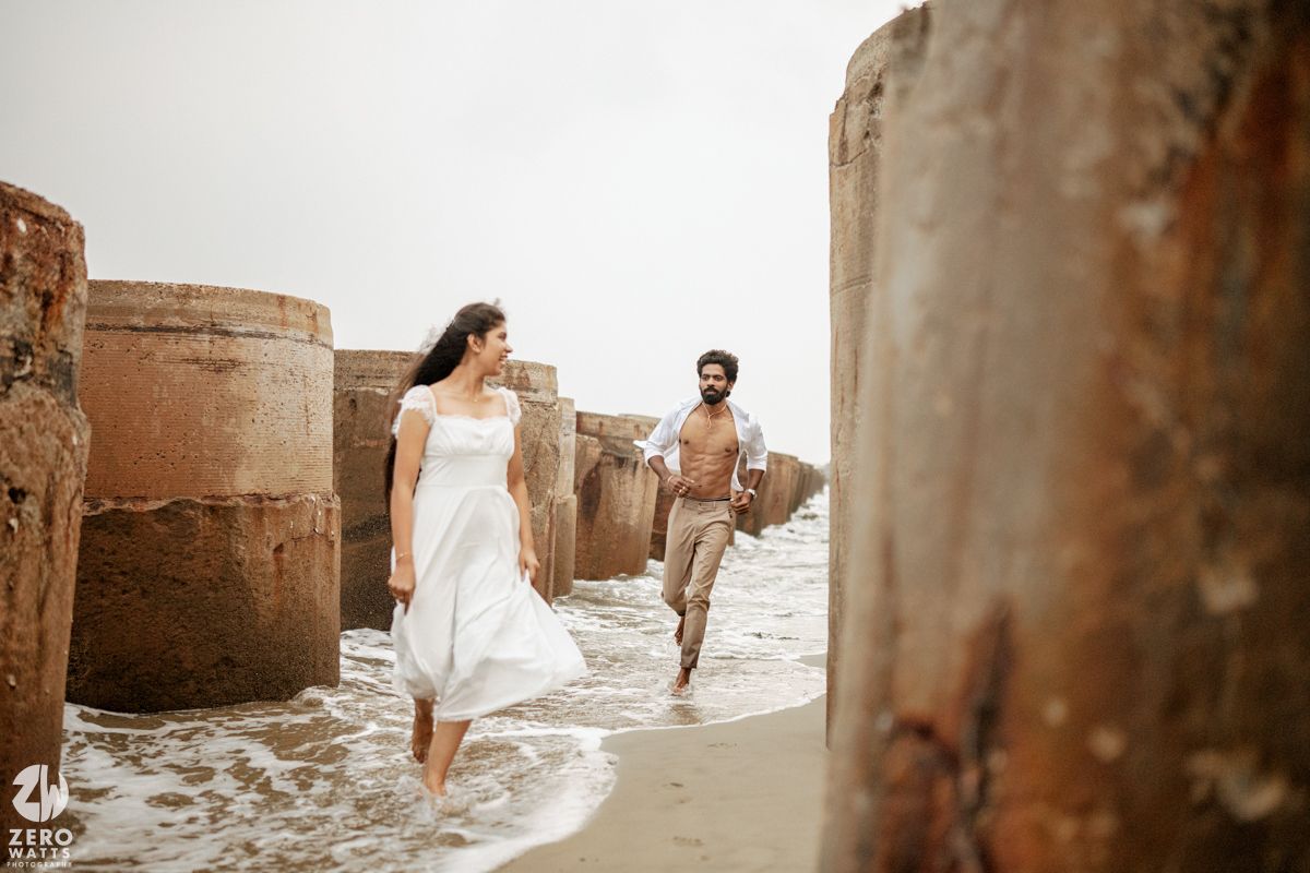 Couple walking along a beach between rustic stone pillars, woman in white dress, man shirtless in tan pants.
