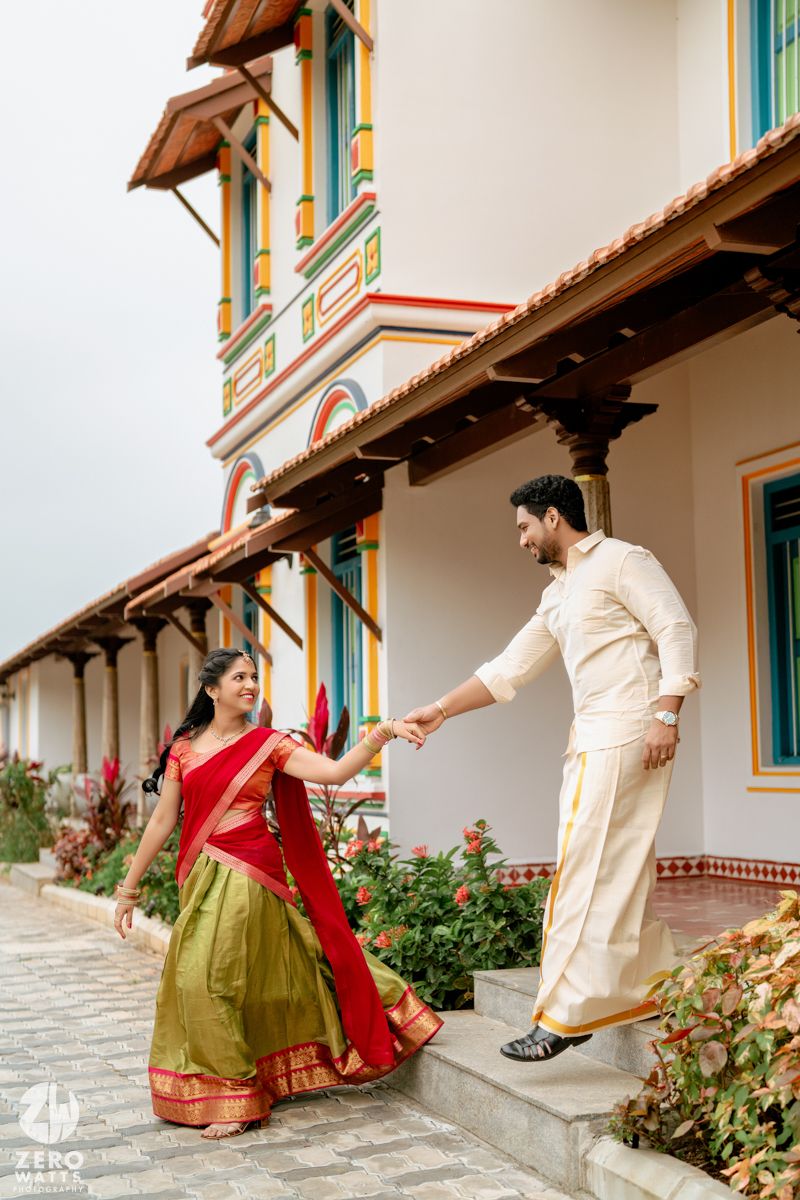 Couple in traditional Indian attire holding hands on temple steps, bride in red and green half saree, groom in white dhoti.