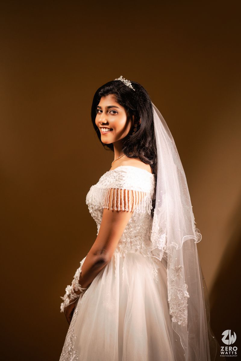 A smiling bride in a white off-shoulder lace gown with veil and tiara poses against a warm brown background.