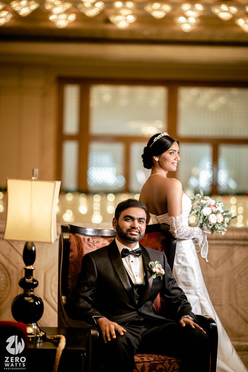 Groom in black tuxedo seated as bride in white gown holds bouquet in ornate ballroom.