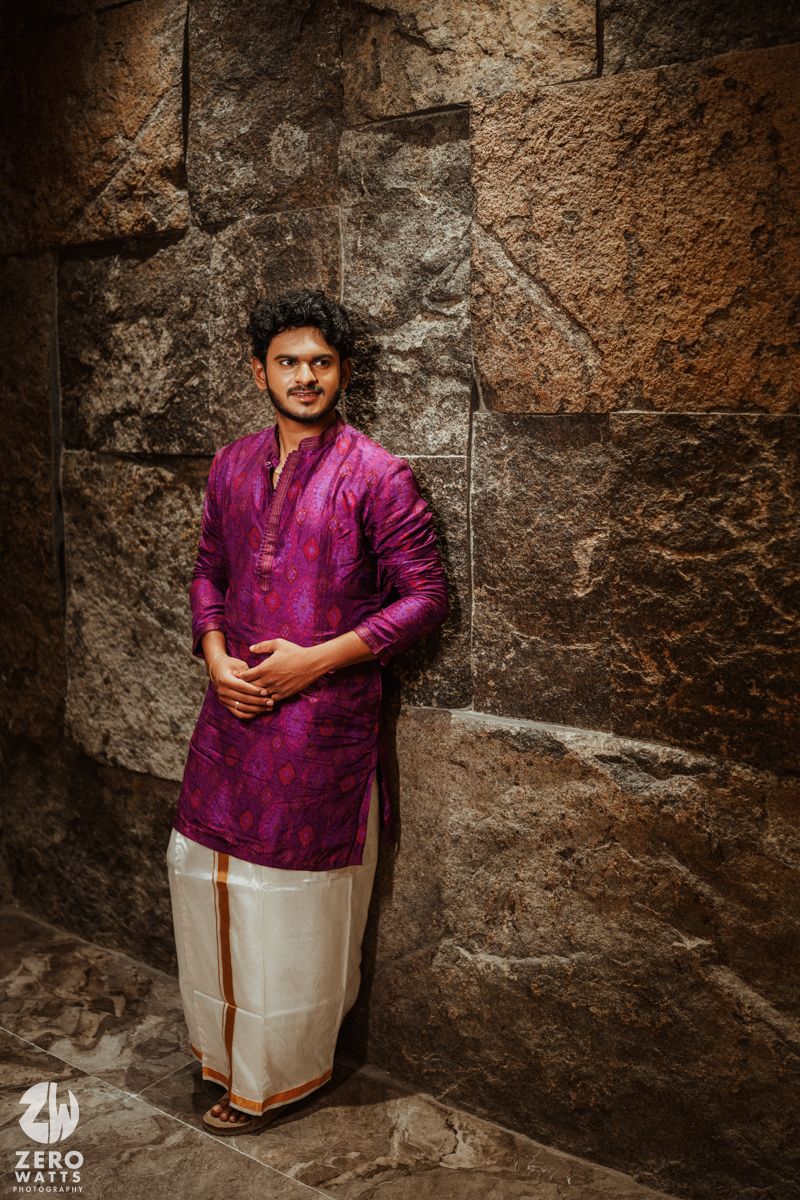 Young man in purple kurta and white dhoti poses against rustic stone wall in traditional attire.