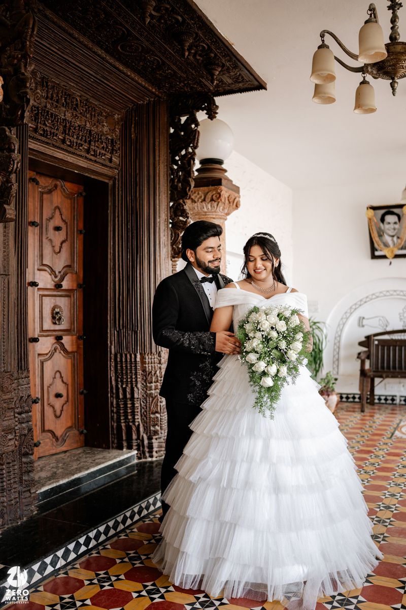 Bride in white tiered gown holding floral bouquet stands with groom in black tuxedo inside ornate wooden doorway.