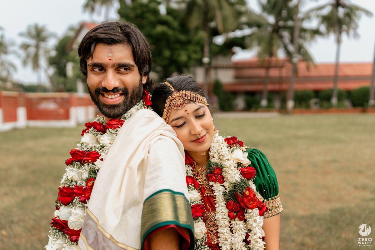 South Indian bride and groom in traditional wedding attire with floral garlands posing outdoors on lush green lawn.