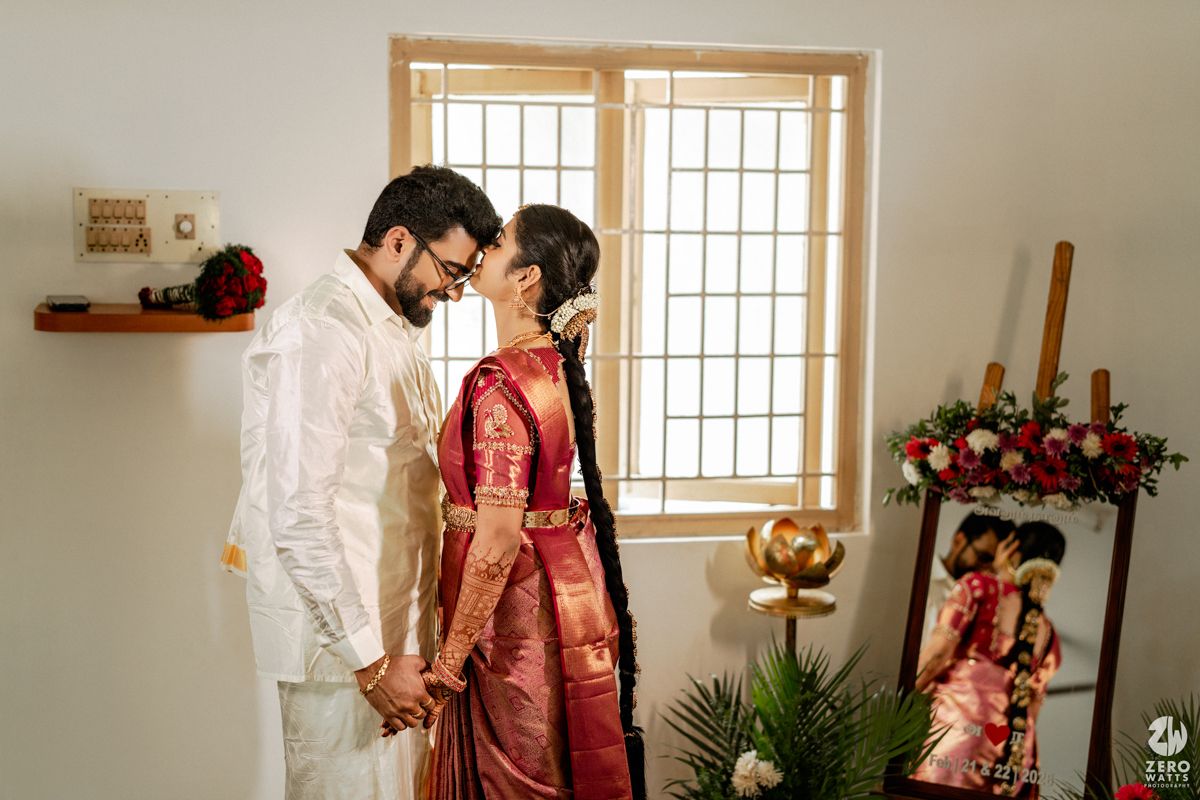 South Indian couple in traditional wedding attire sharing a tender moment by a sunlit window.