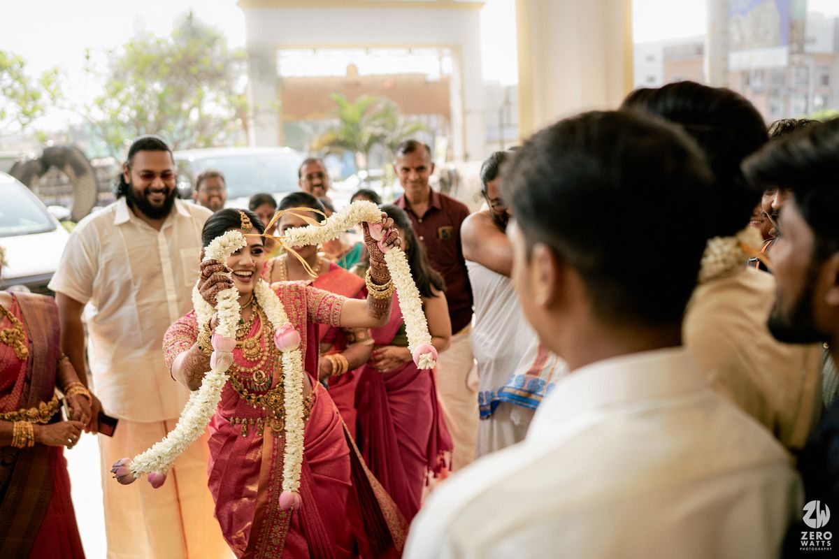 Bride in red saree and gold jewelry walks with garlands during a traditional South Indian wedding ceremony.