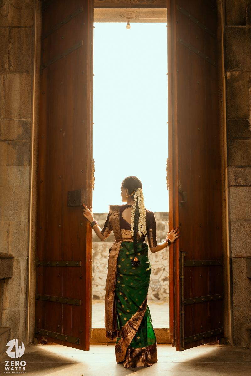 Bride in green Kanjivaram saree stands in ornate temple doorway, hair adorned with flowers, backlit by natural light.