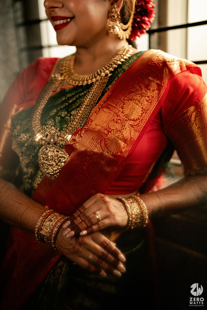 South Indian bride in red Kanjivaram silk saree with gold temple jewelry, mehndi hands, and traditional bridal accessories smiling.