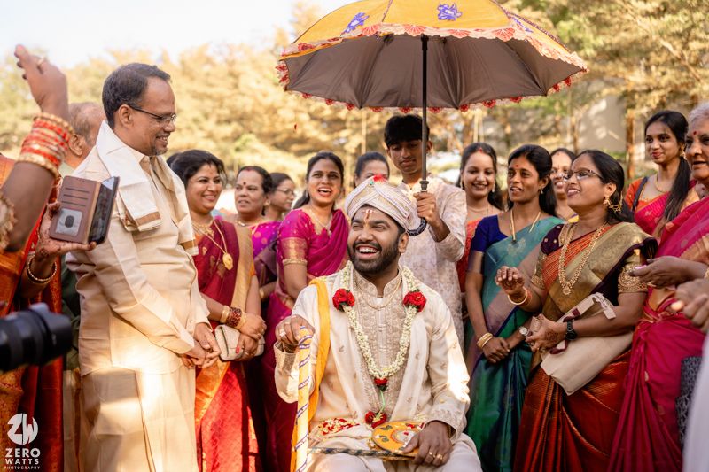 Groom in traditional South Indian wedding attire seated under decorative umbrella surrounded by family during outdoor ceremony.