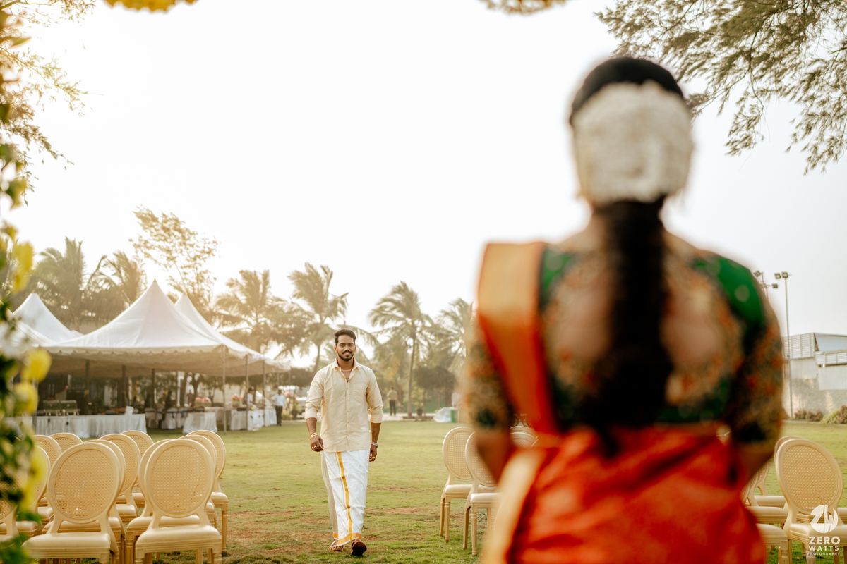 Groom in white walking toward bride in red saree at outdoor South Indian wedding ceremony with floral decorations.