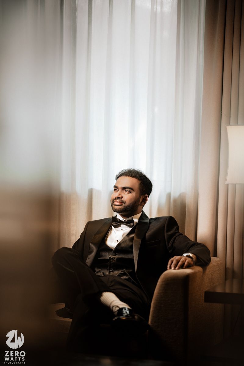 Groom in black tuxedo and bow tie seated in armchair by sheer curtains, smiling elegantly.
