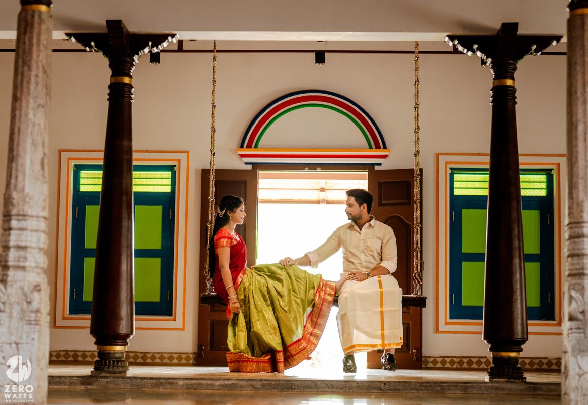 Couple in traditional South Indian attire posing on a wooden swing in a colorfully decorated heritage home.