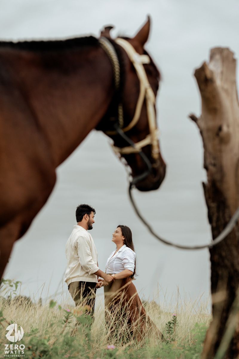 A couple sits together in tall grass with a brown horse standing nearby under cloudy skies.
