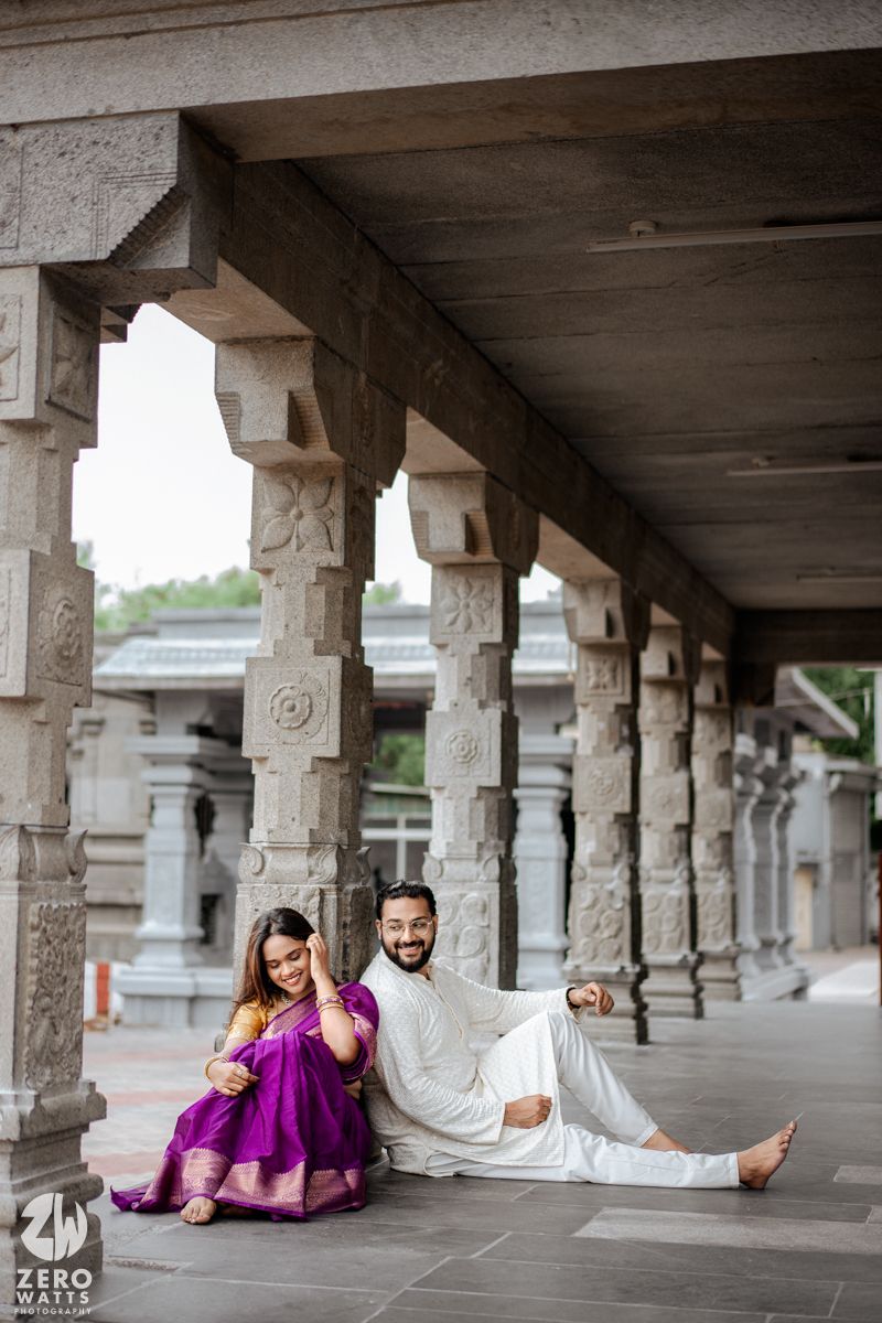Couple posing in traditional Indian attire under a stone colonnade with ornate architectural details.