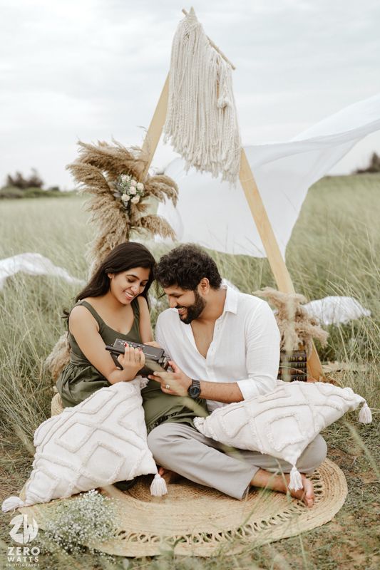 A couple sits on a round rug in a grassy field with a white fabric tent and pampas grass decor for a romantic picnic.