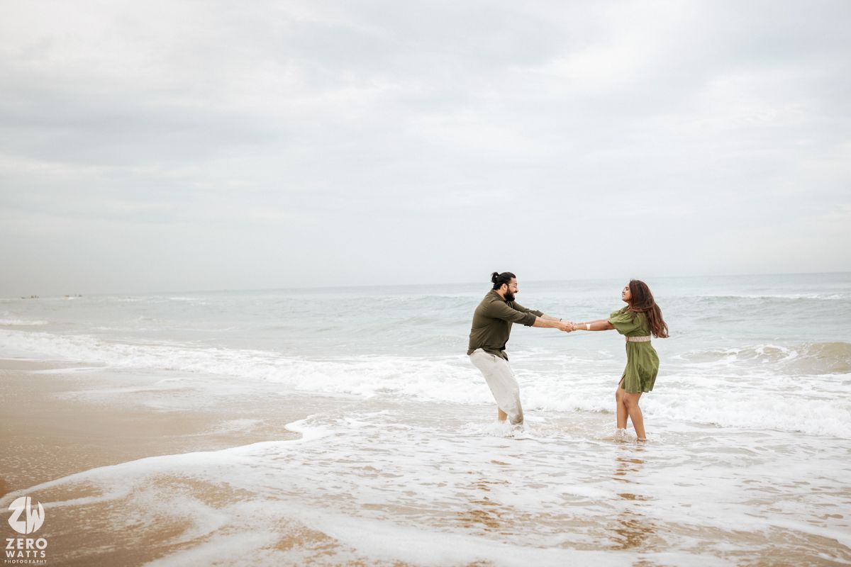 A couple holds hands while playing in the shallow ocean waves on an overcast day at the beach.