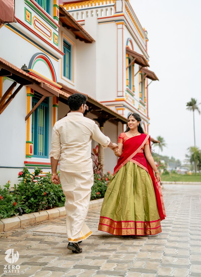 Couple in traditional Indian attire holding hands outside a colorful heritage building, woman in green and red lehenga.
