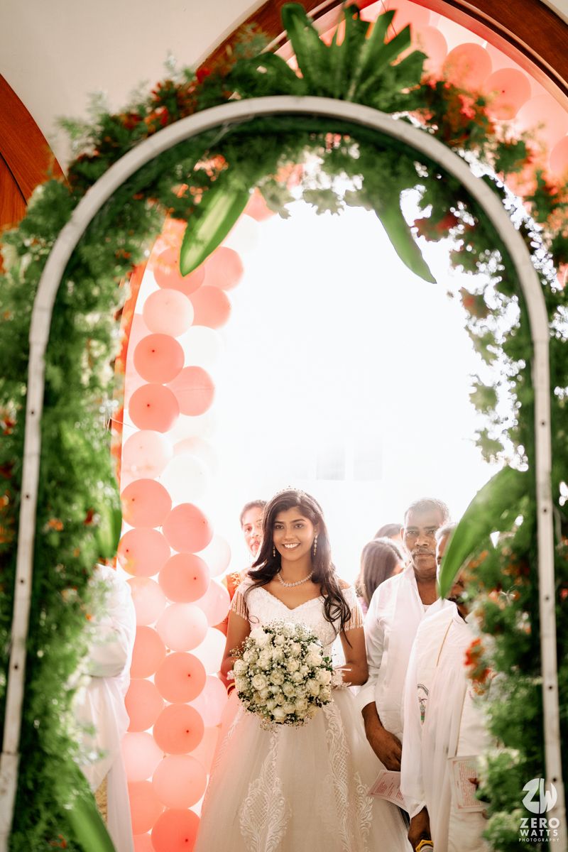 Smiling bride in white gown holding bouquet walks through floral arch with pink balloons and greenery at wedding ceremony.