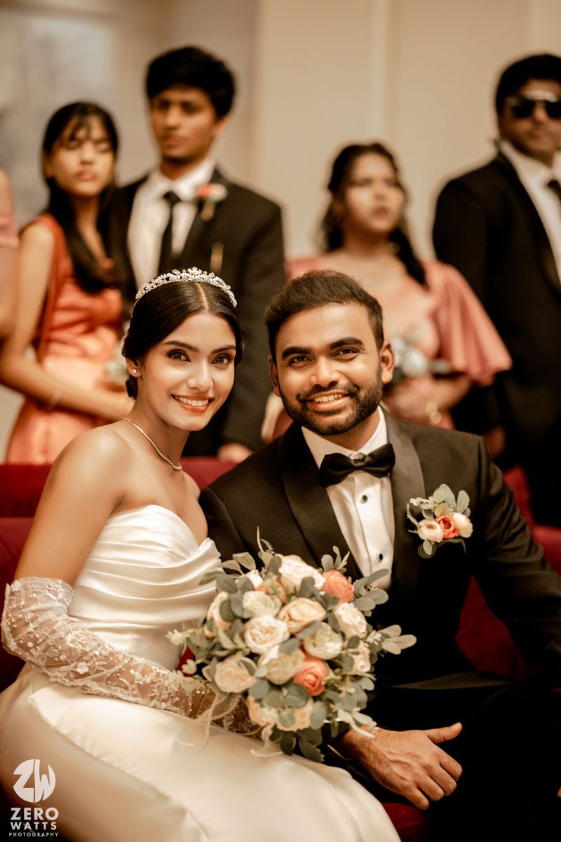 A smiling bride in white gown with tiara holds floral bouquet beside groom in black tuxedo at elegant wedding ceremony.