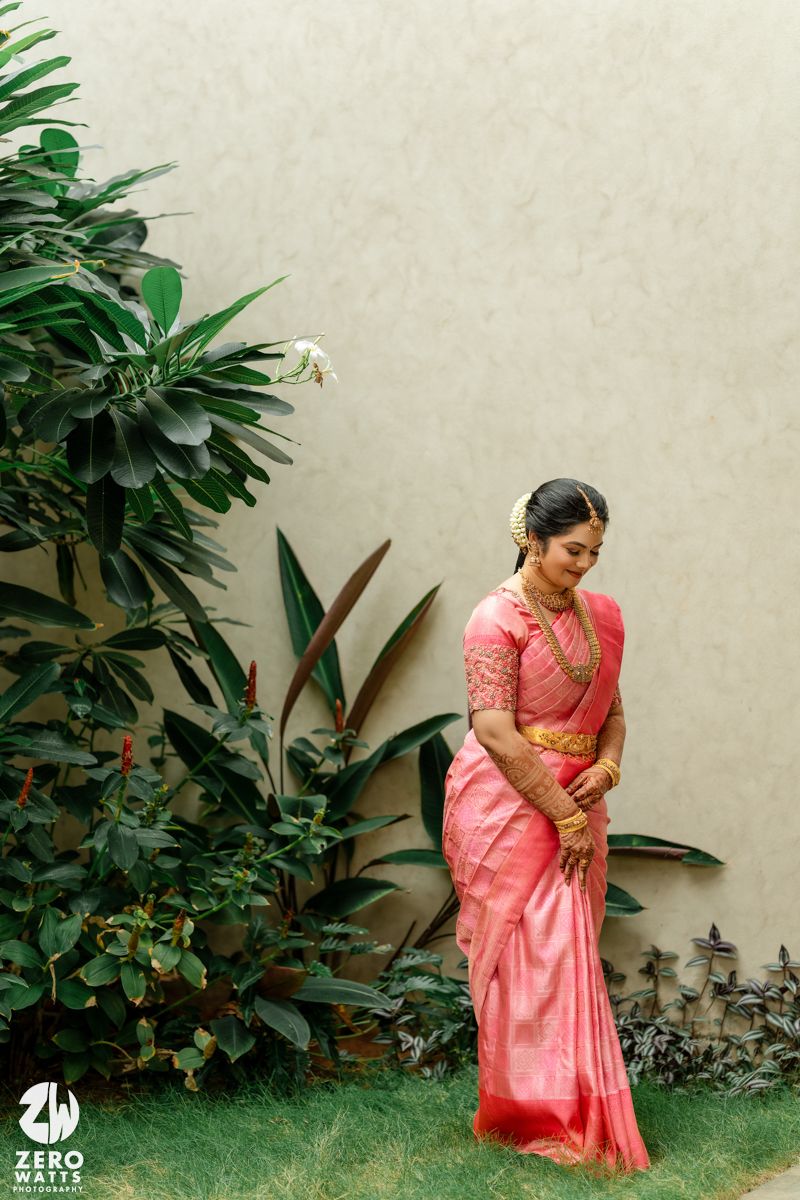 Bride in pink silk saree with gold jewelry poses gracefully outdoors near lush green tropical plants.