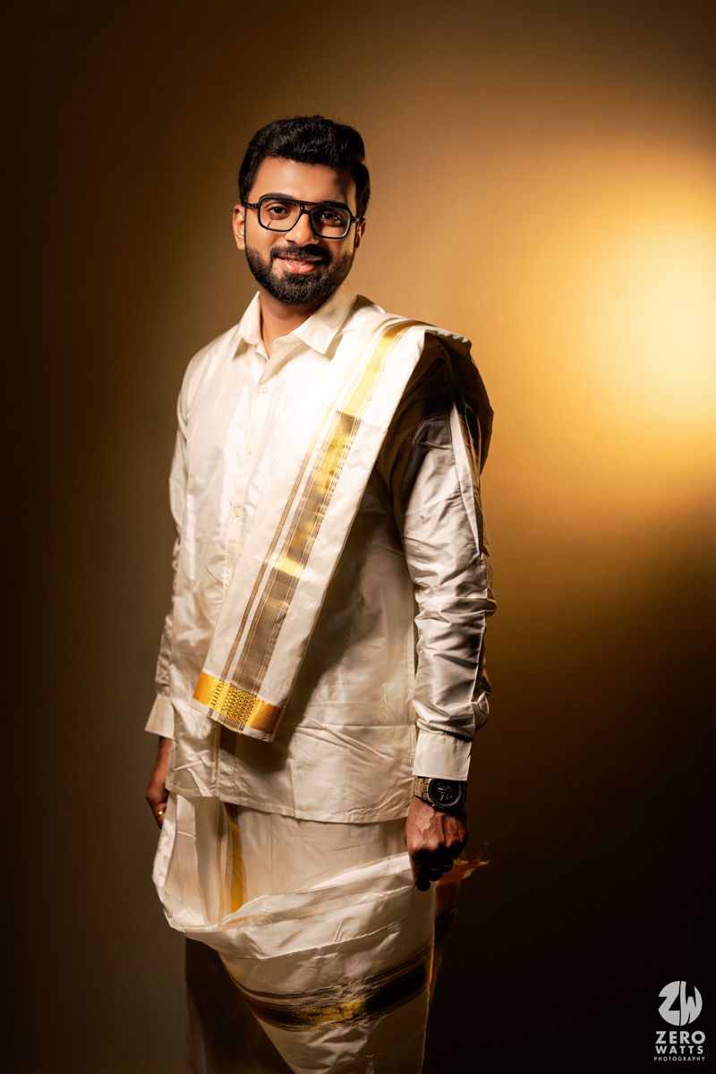 South Indian groom in traditional white silk dhoti with gold border smiles confidently against warm background.