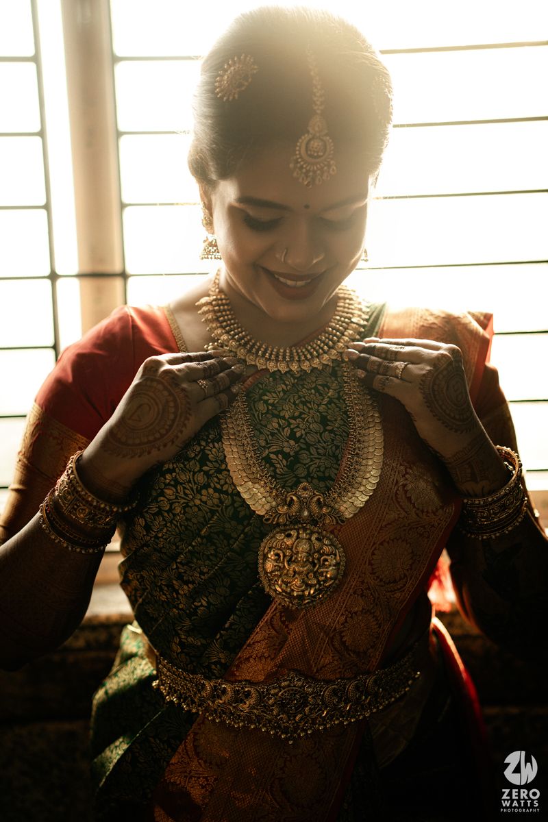 South Indian bride in traditional silk saree adjusting gold temple jewelry necklace, with maang tikka and bangles.