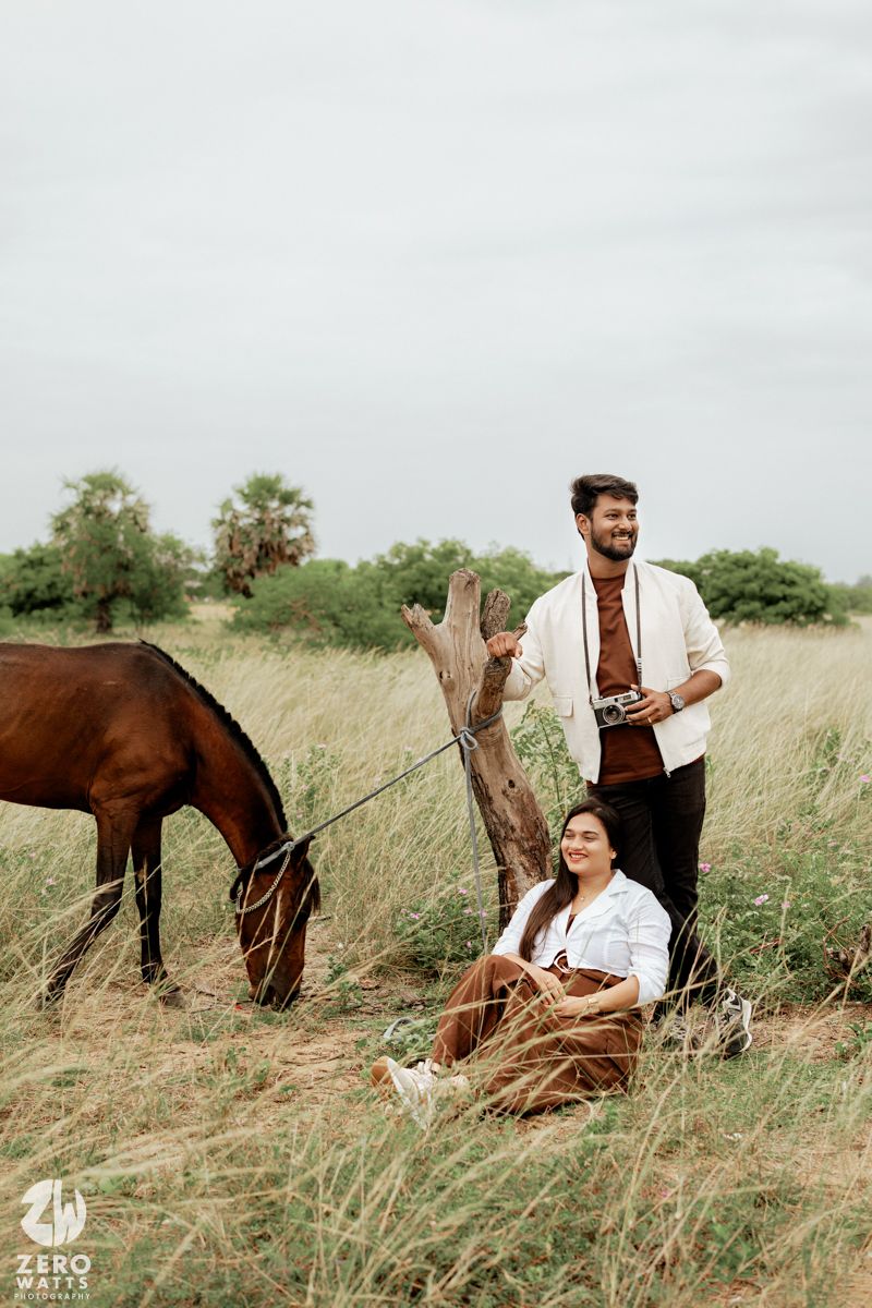 A couple relaxes in tall grass beside their grazing horse during an outdoor photoshoot.