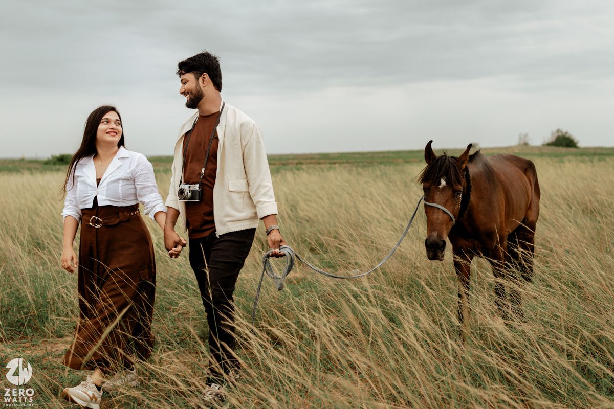 A couple walks together through tall grass with a brown horse following behind them.