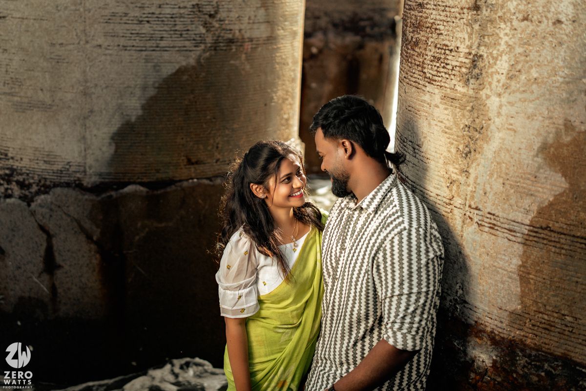 A couple poses playfully against concrete pillars in an urban setting.