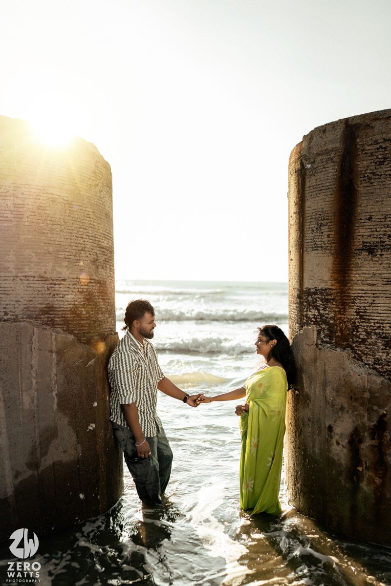 Couple shares intimate moments between concrete pillars as ocean waves crash around them at sunset.