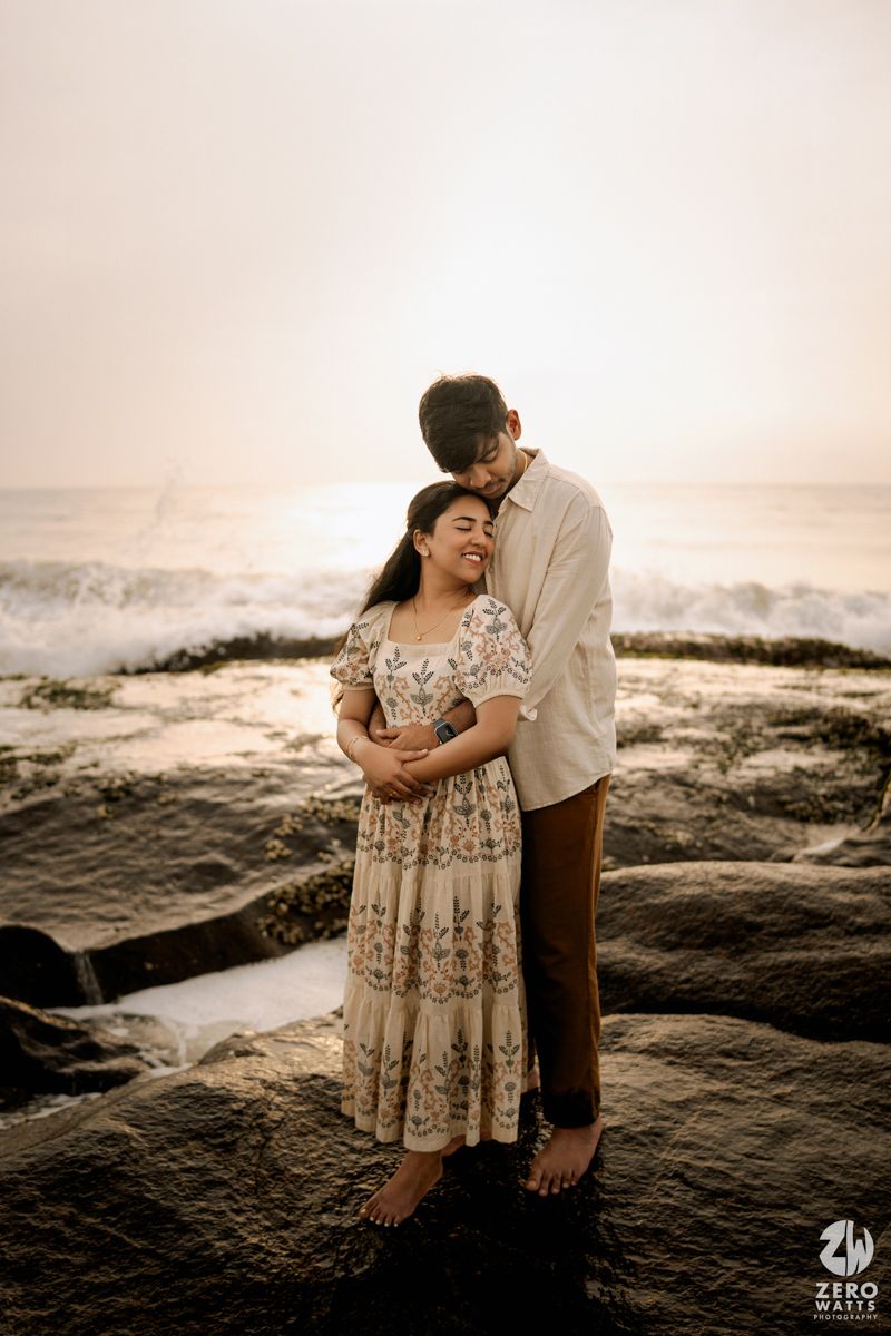 A couple embraces intimately on rocky coastal cliffs at sunset with ocean waves crashing in the background.