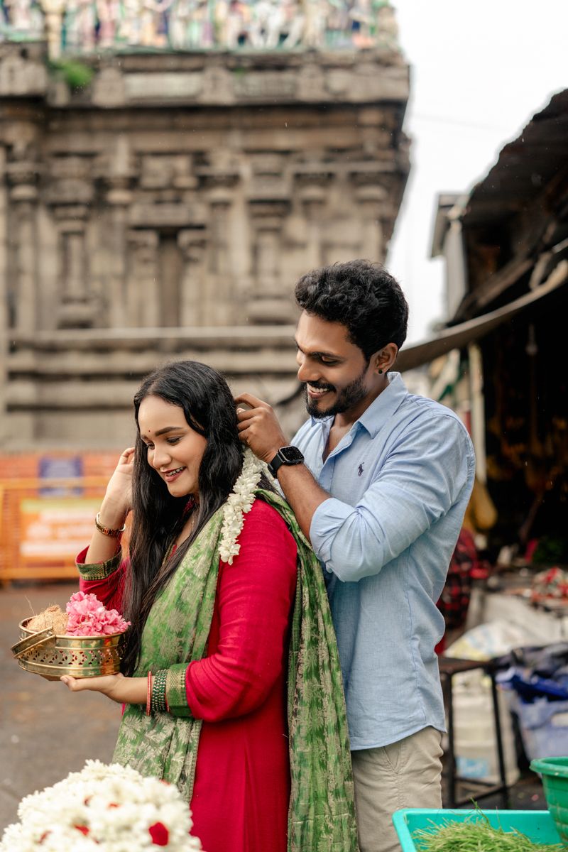 A couple in traditional Indian dress poses with flower petals in front of historic architecture.