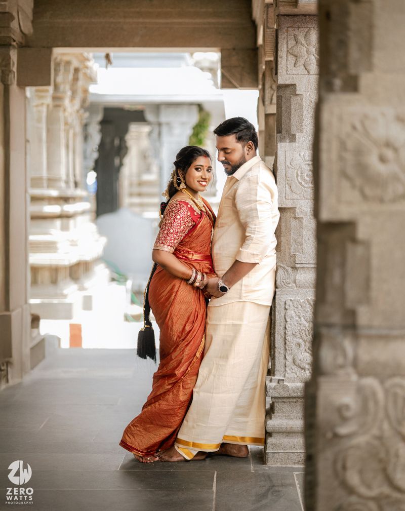 A couple in traditional Indian attire shares romantic moments in a temple corridor with ornate stone pillars.