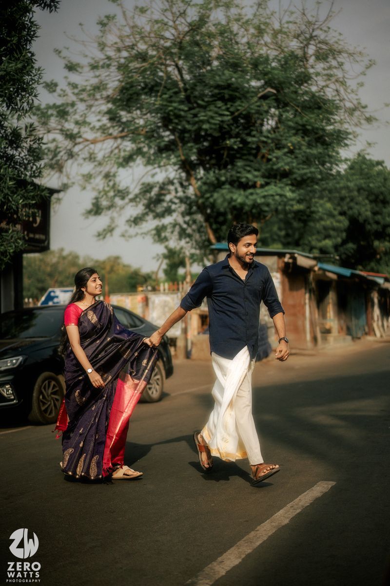 A couple in traditional Indian clothing poses playfully on a street with trees in the background at sunset.