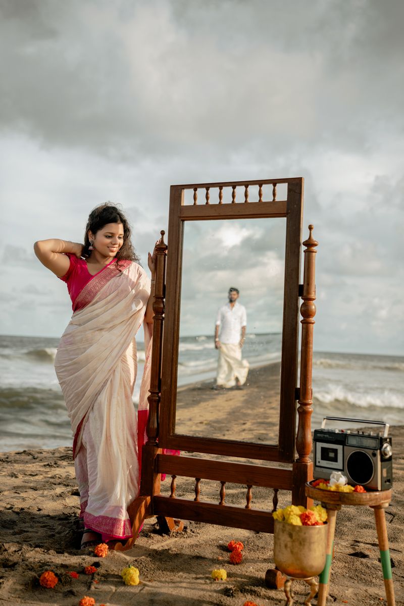 Artistic beach photography scene with a wooden mirror frame and scattered floral decorations against moody skies.