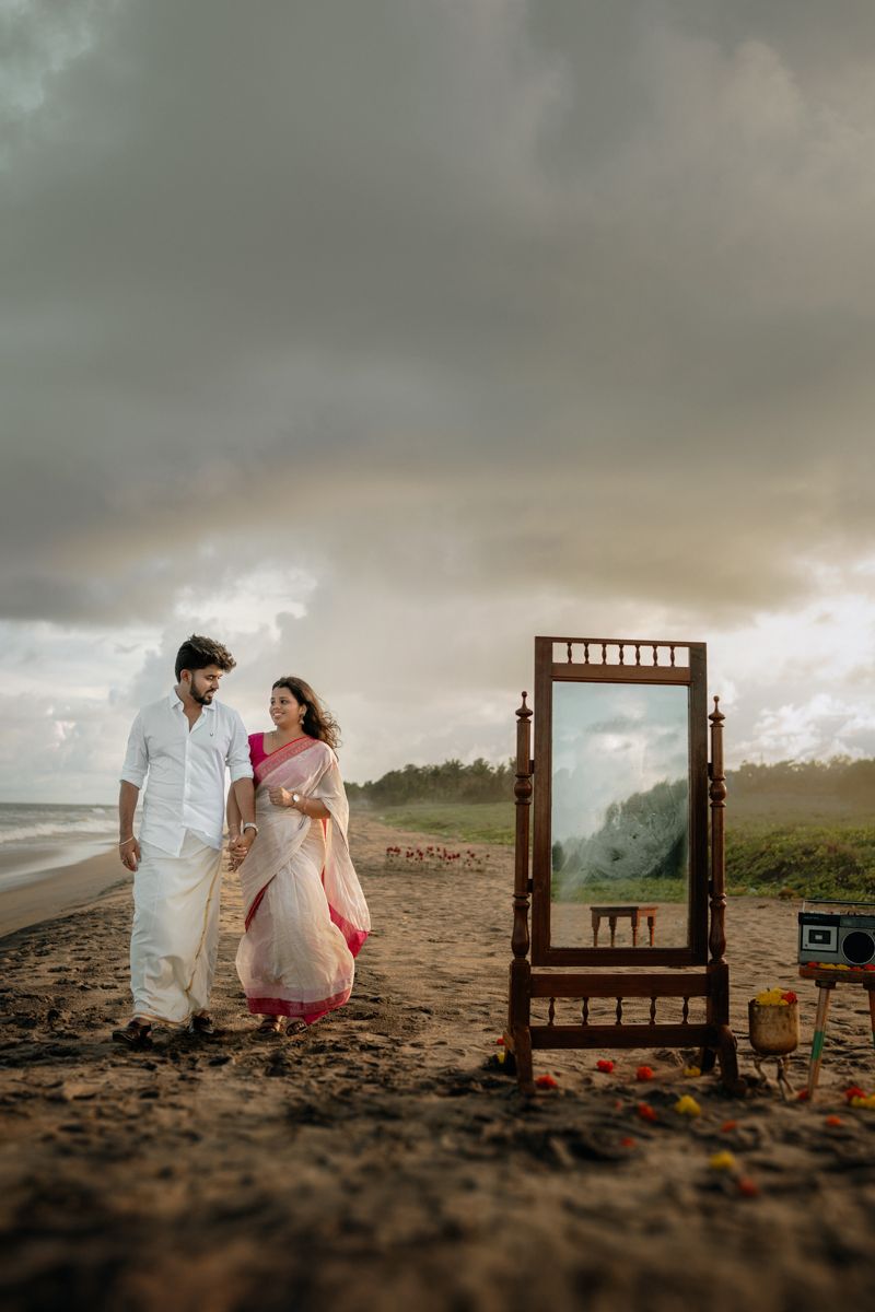 A romantic couple sits on a beach at sunset with an artistic wooden frame installation creating a dreamy atmosphere.