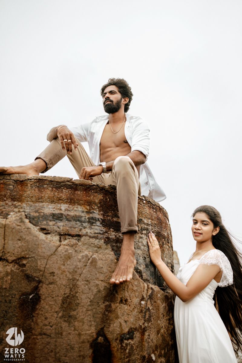 Man sitting atop a rocky structure with open white shirt, woman in white dress standing below, beach backdrop.