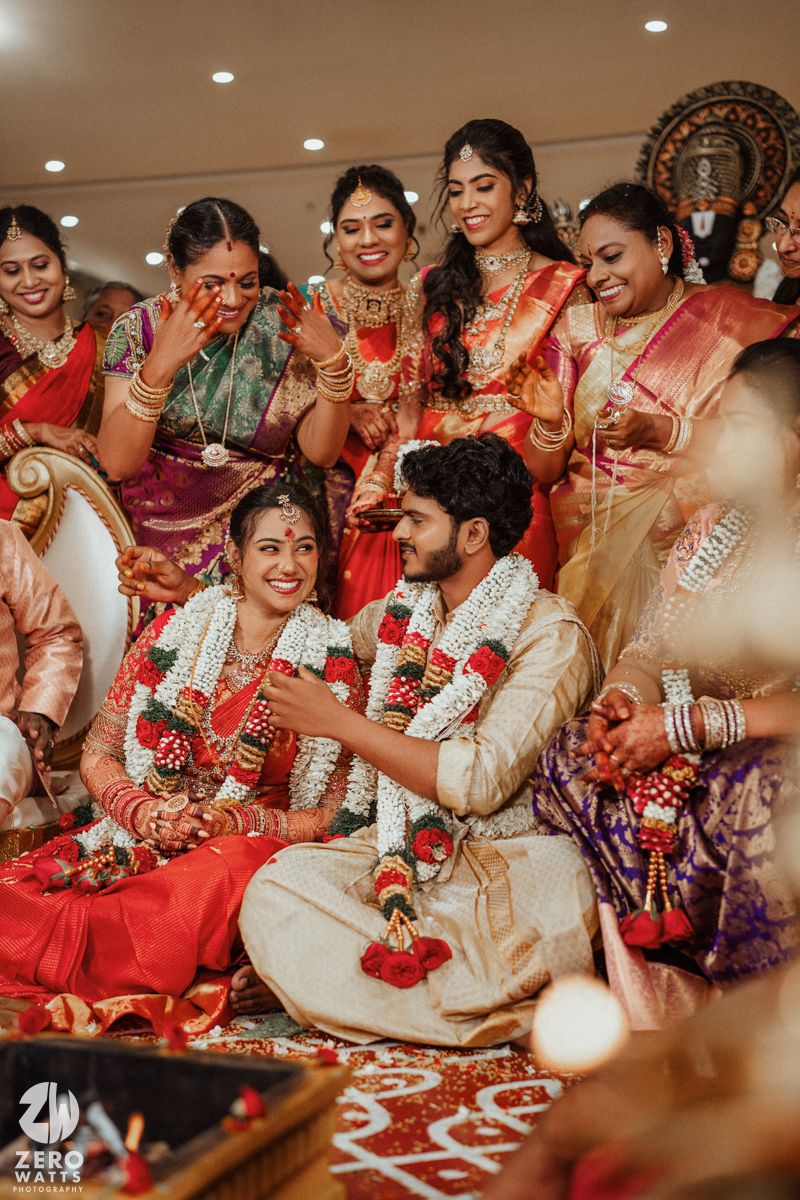 South Indian wedding couple in red and gold attire surrounded by joyful bridesmaids during traditional ceremony.