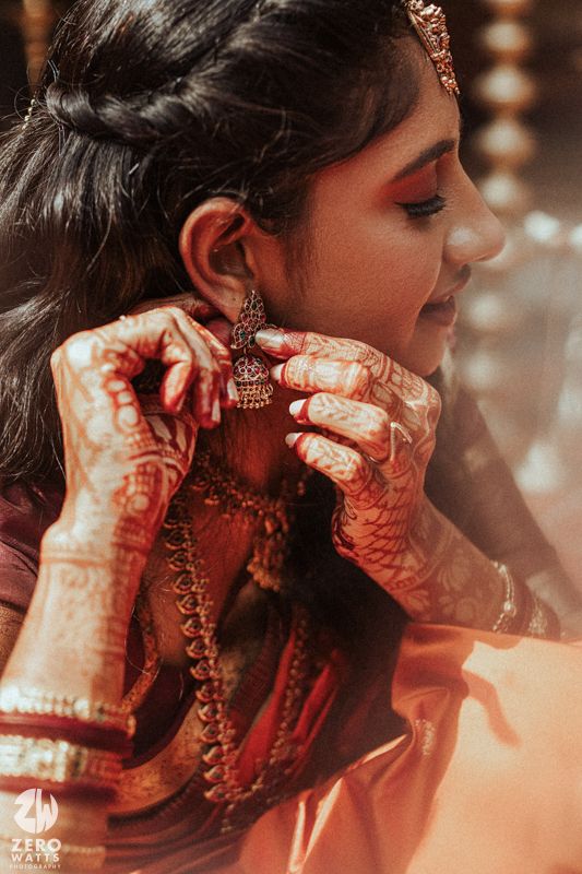 Indian bride with mehndi-covered hands adjusting traditional jhumka earring, wearing red saree and gold jewelry.
