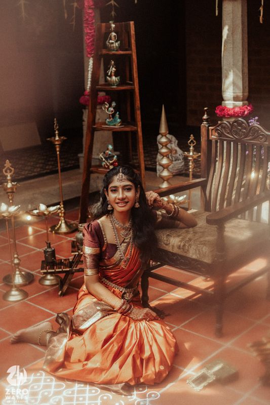 Young woman in orange and maroon silk saree with gold jewelry sits gracefully on tiled floor amid brass lamps.