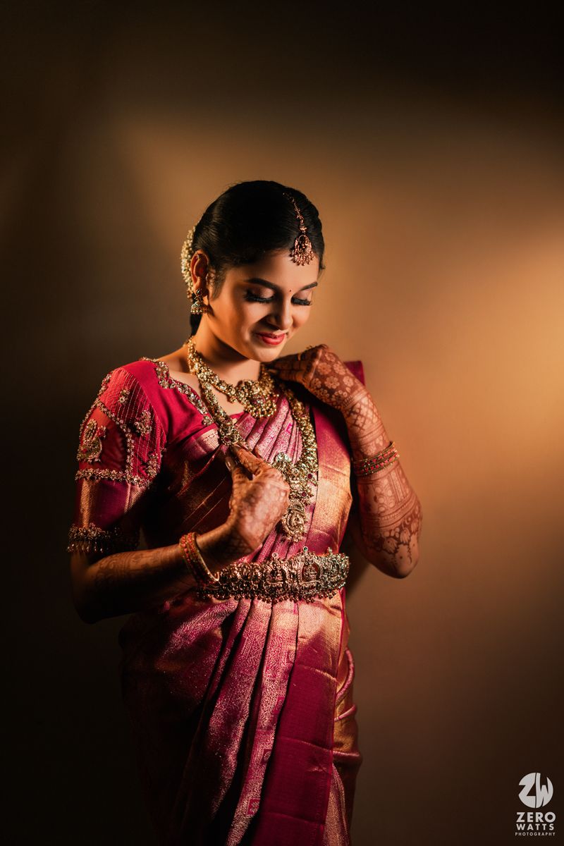 South Indian bride in pink silk saree with gold jewelry and mehndi poses gracefully against warm background.